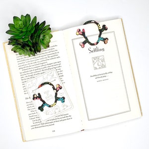 May include: A colourful bookmark shaped like a skull with the words "Read in Peace" on it. The bookmark is on a page of a book that says "Settling" and has a quote by Robert Bridges.
