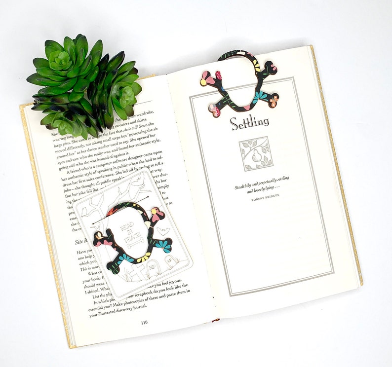 May include: A colourful bookmark shaped like a skull with the words "Read in Peace" on it. The bookmark is on a page of a book that says "Settling" and has a quote by Robert Bridges.