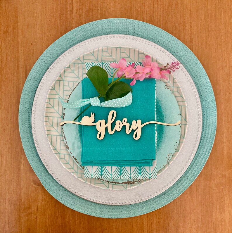 May include: A white and teal layered plate with a teal napkin and a wooden place card that says "glory" with a bunny rabbit silhouette. Pink flowers are on the napkin.