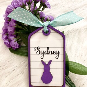 May include: A purple wooden tag with a white background and a purple bunny silhouette. The tag has the name "Sydney" written in black cursive. The tag is tied with a light blue ribbon with white polka dots.