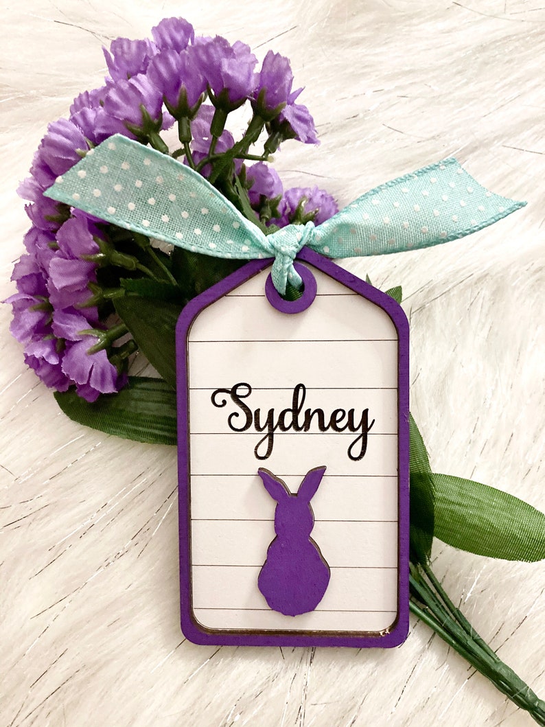 May include: A purple wooden tag with a white background and a purple bunny silhouette. The tag has the name "Sydney" written in black cursive. The tag is tied with a light blue ribbon with white polka dots.