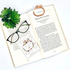 May include: A wooden bookmark with an autumnal design. The bookmark features a tree with leaves in shades of orange, red, and yellow. The text "Carve out time to read" is written on the bookmark. The bookmark is resting on an open book with a pair of glasses.