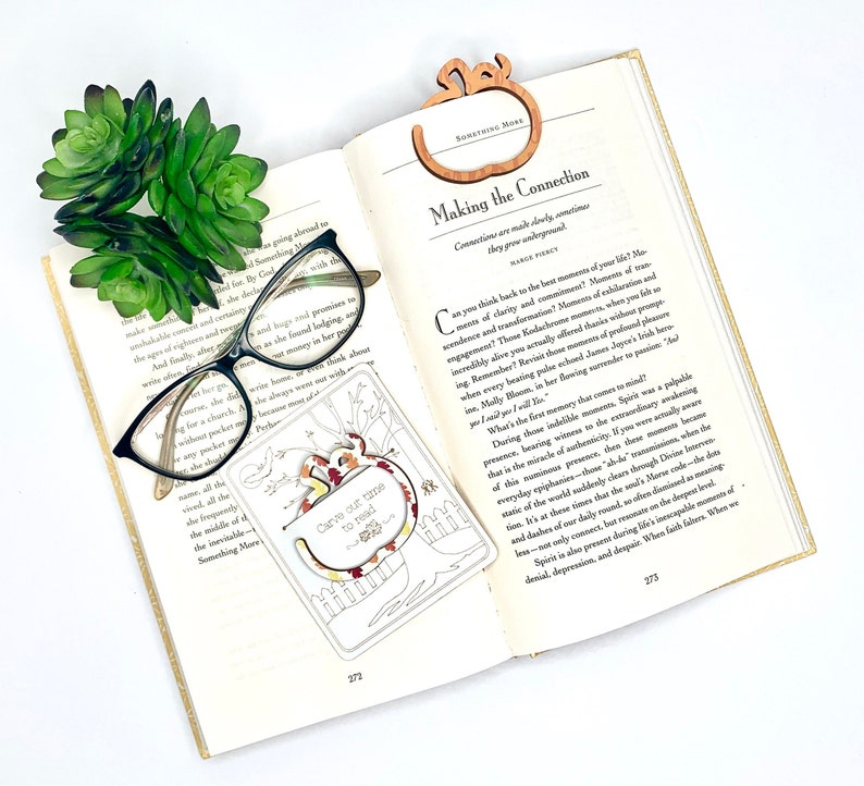 May include: A wooden bookmark with an autumnal design. The bookmark features a tree with leaves in shades of orange, red, and yellow. The text "Carve out time to read" is written on the bookmark. The bookmark is resting on an open book with a pair of glasses.