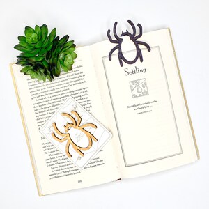 May include: A close-up of an open book with a wooden spider bookmark. The book is open to a page with the title "Settling" and a quote by Robert Bridges. The bookmark is a light brown colour and has a spider web design. The book is on a white surface.