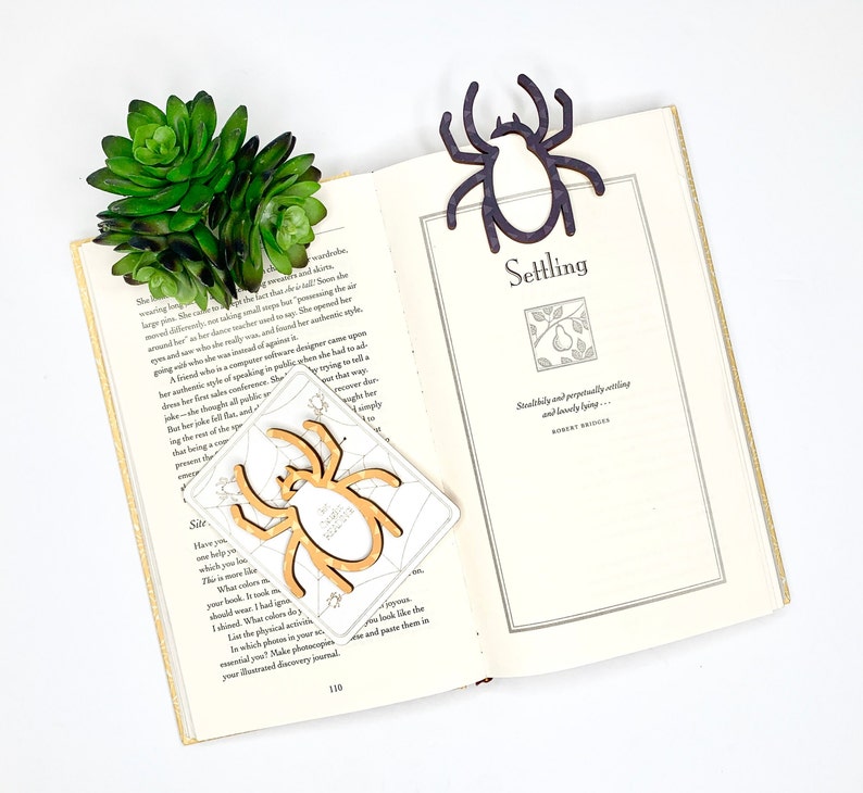 May include: A close-up of an open book with a wooden spider bookmark. The book is open to a page with the title "Settling" and a quote by Robert Bridges. The bookmark is a light brown colour and has a spider web design. The book is on a white surface.