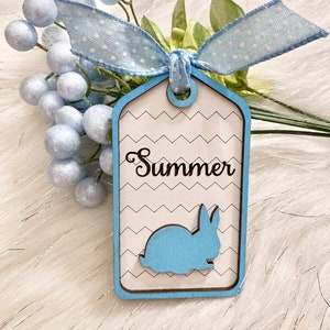 May include: A blue and white wooden tag with a white chevron pattern. The tag has a blue bunny silhouette and the word "Summer" written in black cursive.