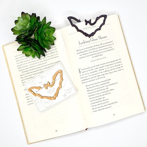 May include: A wooden bat bookmark with the words "Batty for Books" printed on it. The bookmark is on an open book with a green succulent plant on top. The book is open to a page with the title "Looking Glass Shame" and a poem by George Bernard Shaw.