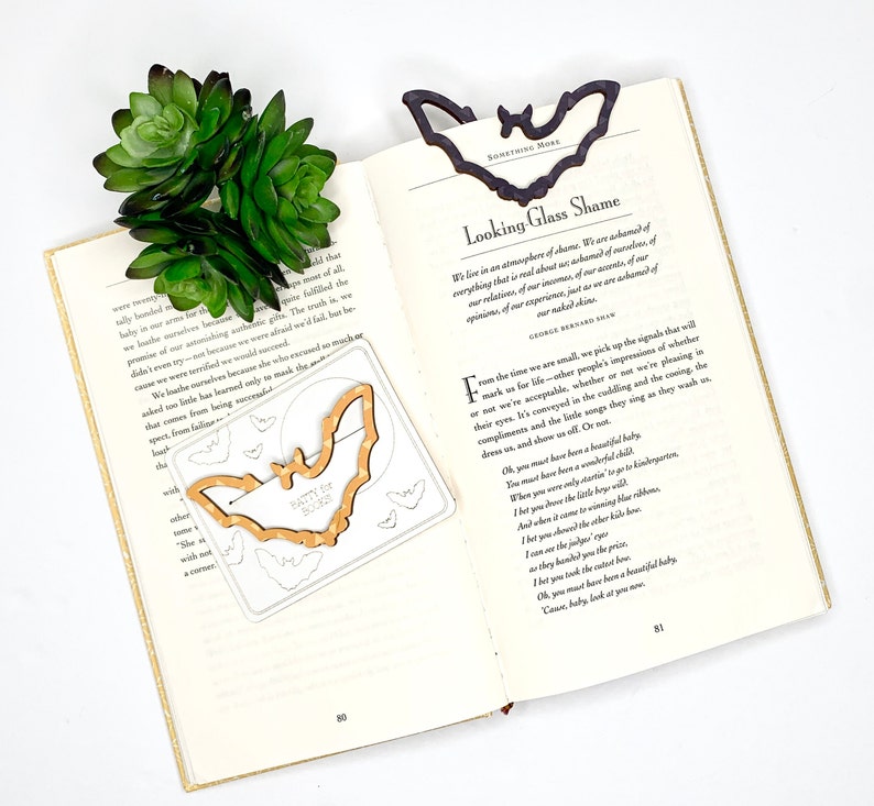 May include: A wooden bat bookmark with the words "Batty for Books" printed on it. The bookmark is on an open book with a green succulent plant on top. The book is open to a page with the title "Looking Glass Shame" and a poem by George Bernard Shaw.