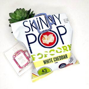 May include: A bag of Skinny Pop popcorn with white cheddar flavour. The bag is yellow and white with blue and red accents. The bag is open and the popcorn is visible. The bag says "Skinny Pop Popcorn White Cheddar Flavour" and "43 Calories".