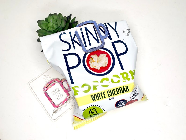 May include: A bag of Skinny Pop popcorn with white cheddar flavour. The bag is yellow and white with blue and red accents. The bag is open and the popcorn is visible. The bag says "Skinny Pop Popcorn White Cheddar Flavour" and "43 Calories".