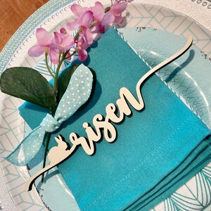 May include: A blue napkin with a white polka dot ribbon and a sprig of pink flowers. A wooden place card with the word "risen" is attached to the napkin.