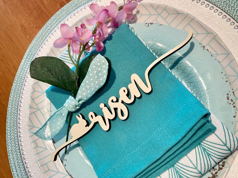 May include: A blue napkin with a white polka dot ribbon and a sprig of pink flowers. A wooden place card with the word "risen" is attached to the napkin.