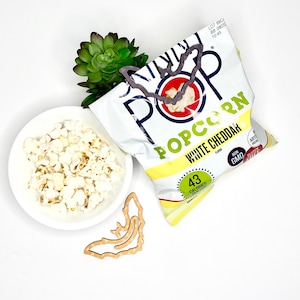 May include: A bag of white cheddar popcorn with a bat-shaped cutout. The bag is yellow and white with the words "Skinny Pop Popcorn White Cheddar" printed on it. The bag is sitting on a white surface with a small bowl of popcorn and a wooden bat cutout.