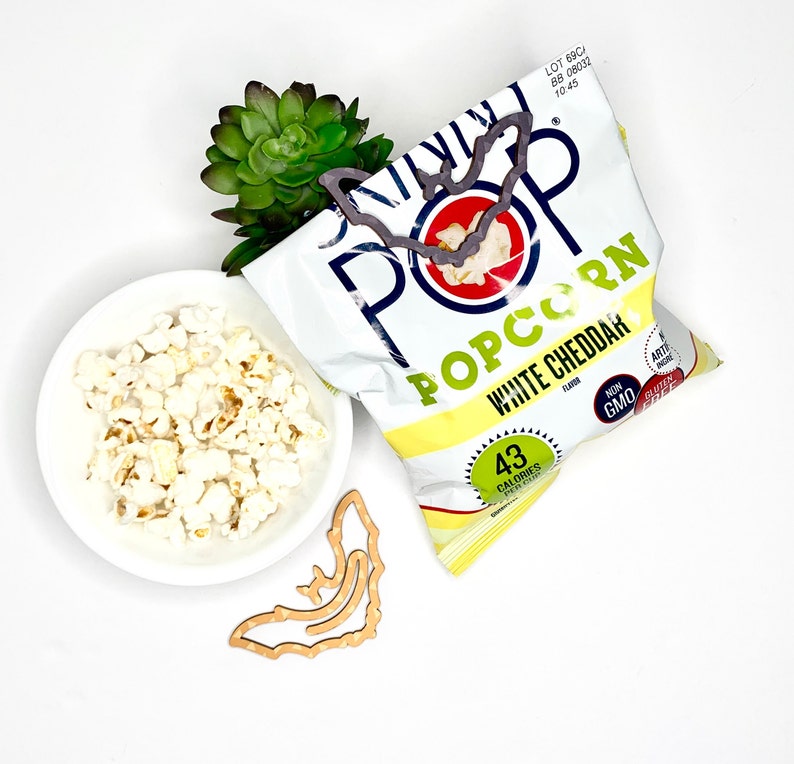 May include: A bag of white cheddar popcorn with a bat-shaped cutout. The bag is yellow and white with the words "Skinny Pop Popcorn White Cheddar" printed on it. The bag is sitting on a white surface with a small bowl of popcorn and a wooden bat cutout.