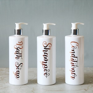 Puede incluir: Tres botellas de bomba blancas con tapas plateadas. Cada botella tiene una etiqueta escrita en color cobre: "Bath Soap", "Shampoo" y "Conditioner". Las botellas están dispuestas sobre una superficie clara contra un fondo azul pálido.