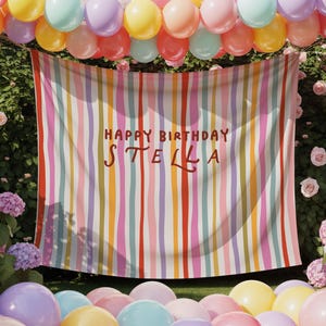 May include: A colorful birthday party scene with a striped backdrop that reads "Happy Birthday Stella." Balloons in various colors hang above the backdrop and are scattered on the ground. Pink, purple, and yellow flowers surround the scene.