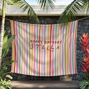 May include: A colorful striped banner with the text "HAPPY BIRTHDAY STELLA" in brown lettering. The banner features vertical stripes in various colors, including pink, orange, yellow, and green. The banner is hanging outdoors.