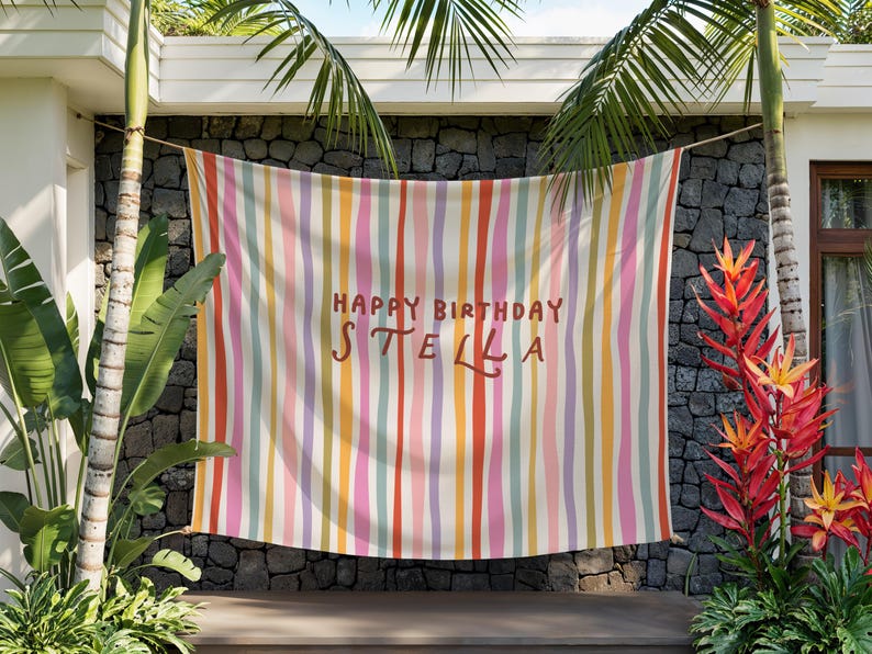 May include: A colorful striped banner with the text "HAPPY BIRTHDAY STELLA" in brown lettering. The banner features vertical stripes in various colors, including pink, orange, yellow, and green. The banner is hanging outdoors.