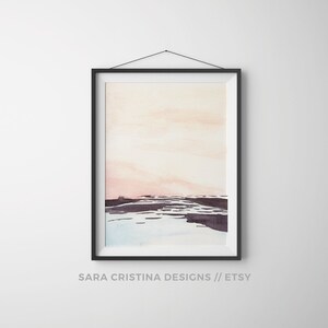 Peut inclure: Une peinture &agrave; l'aquarelle encadr&eacute;e d'un paysage avec un ciel rose et orange et des collines violettes fonc&eacute;es au premier plan. La peinture est dans un cadre noir et est accroch&eacute;e &agrave; un mur blanc.