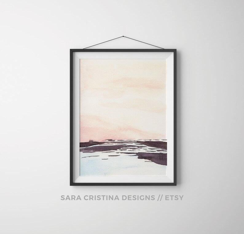 Peut inclure: Une peinture &agrave; l'aquarelle encadr&eacute;e d'un paysage avec un ciel rose et orange et des collines violettes fonc&eacute;es au premier plan. La peinture est dans un cadre noir et est accroch&eacute;e &agrave; un mur blanc.