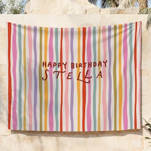 May include: A colorful banner with vertical stripes in red, orange, yellow, green, blue, pink, and purple. The words "HAPPY BIRTHDAY STELLA" are written in brown script across the center of the banner. The banner is hanging on a beige wall.