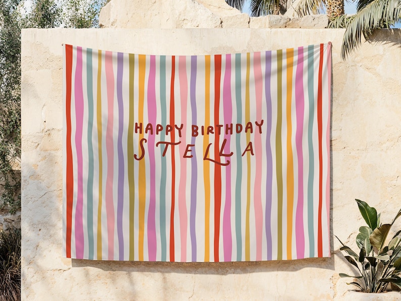 May include: A colorful banner with vertical stripes in red, orange, yellow, green, blue, pink, and purple. The words "HAPPY BIRTHDAY STELLA" are written in brown script across the center of the banner. The banner is hanging on a beige wall.