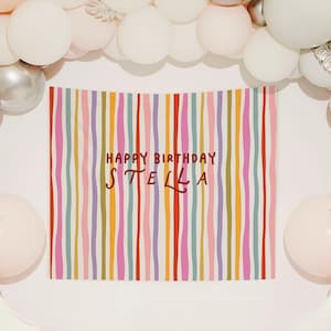 May include: A colorful birthday banner with vertical stripes in various colors, including red, pink, and purple, with the text "HAPPY BIRTHDAY STELLA". The banner is surrounded by white, silver, and pink balloons, creating a festive party atmosphere.