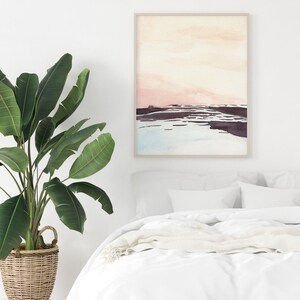 Peut inclure: Une peinture &agrave; l'aquarelle d'un paysage avec un ciel rose et orange, une &eacute;tendue d'eau bleue et une terre brune. La peinture est encadr&eacute;e dans un cadre en bois clair et est accroch&eacute;e &agrave; un mur blanc au-dessus d'un lit blanc avec une couverture blanche.