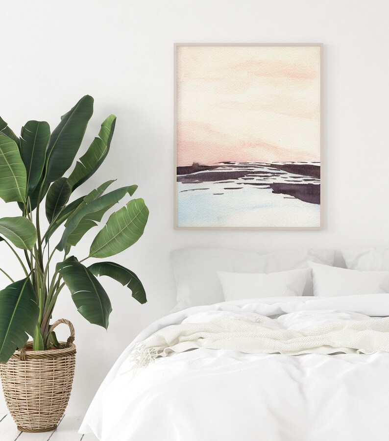 Peut inclure: Une peinture &agrave; l'aquarelle d'un paysage avec un ciel rose et orange, une &eacute;tendue d'eau bleue et une terre brune. La peinture est encadr&eacute;e dans un cadre en bois clair et est accroch&eacute;e &agrave; un mur blanc au-dessus d'un lit blanc avec une couverture blanche.