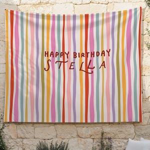 May include: A colorful wall hanging with vertical stripes in shades of red, pink, orange, yellow, green, and blue. The words "HAPPY BIRTHDAY STELLA" are written in brown script across the center of the fabric.