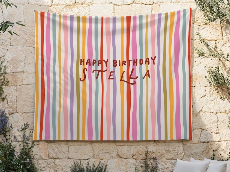 May include: A colorful wall hanging with vertical stripes in shades of red, pink, orange, yellow, green, and blue. The words "HAPPY BIRTHDAY STELLA" are written in brown script across the center of the fabric.