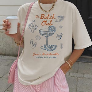 May include: Cream-colored t-shirt with a blue and orange graphic that reads "the Batch Club" and "Jess's Bachelorette, Lincoln City, Oregon." The shirt features illustrations of beach-themed items. The person is wearing pink striped shorts.