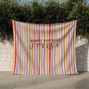 May include: A colorful banner with vertical stripes in shades of red, pink, yellow, orange, and purple. The words "HAPPY BIRTHDAY STELLA" are written in a handwritten style in the center of the banner. The banner is hanging outside.