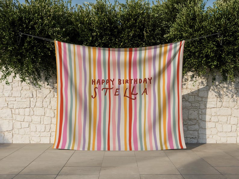 May include: A colorful banner with vertical stripes in shades of red, pink, yellow, orange, and purple. The words "HAPPY BIRTHDAY STELLA" are written in a handwritten style in the center of the banner. The banner is hanging outside.