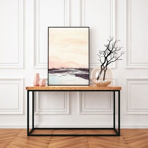 Peut inclure: Une peinture abstraite aquarelle de paysage rose et bleu encadr&eacute;e de noir est suspendue au-dessus d'une console en bois avec un cadre en m&eacute;tal noir. Un vase en verre transparent avec des pierres blanches et une branche s&eacute;ch&eacute;e est pos&eacute; sur la table.