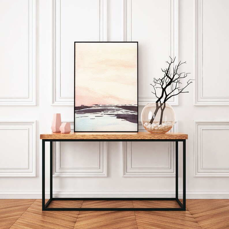 Peut inclure: Une peinture abstraite aquarelle de paysage rose et bleu encadr&eacute;e de noir est suspendue au-dessus d'une console en bois avec un cadre en m&eacute;tal noir. Un vase en verre transparent avec des pierres blanches et une branche s&eacute;ch&eacute;e est pos&eacute; sur la table.