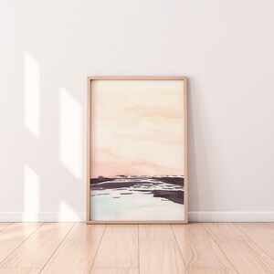 Peut inclure: Une peinture &agrave; l'aquarelle encadr&eacute;e d'un paysage avec un ciel rose p&acirc;le et orange et un premier plan bleu fonc&eacute; et gris. La peinture est dans un cadre en bois clair.