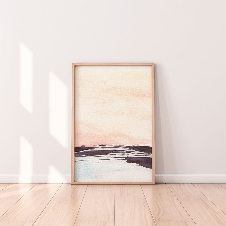 Peut inclure: Une peinture &agrave; l'aquarelle encadr&eacute;e d'un paysage avec un ciel rose p&acirc;le et orange et un premier plan bleu fonc&eacute; et gris. La peinture est dans un cadre en bois clair.