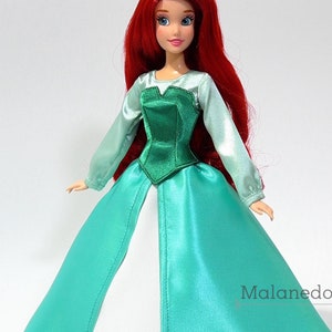 May include: A red-haired doll wearing a teal and green dress with a white underskirt. The doll has blue eyes and is wearing a crown.