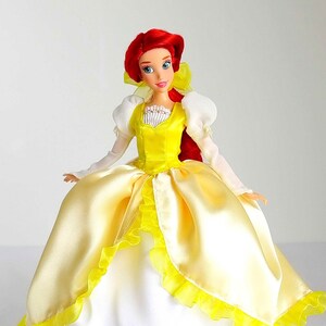 Pode incluir: Uma boneca de cabelo ruivo vestindo um vestido de cetim amarelo com acabamento de renda branca e um corpete branco. A boneca tem olhos azuis e um la&ccedil;o vermelho no cabelo.