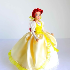 Pode incluir: Uma boneca de cabelo ruivo vestindo um vestido de cetim amarelo com uma borda de renda branca e uma fita amarela no cabelo.
