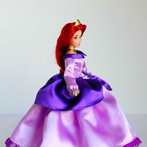 May include: A red-haired doll wearing a purple and pink dress with a ruffled hem. The doll is wearing a gold crown with green gems.