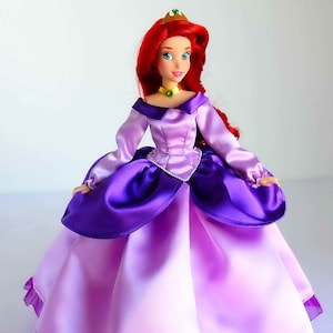 May include: A red-haired doll wearing a purple and pink satin dress with a gold crown. The doll has blue eyes and is wearing a green necklace.