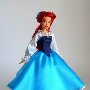 May include: A red-haired doll wearing a blue satin dress with a white lace trim and a navy blue bodice. The doll has blue eyes and is wearing a blue bow in her hair.