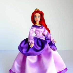 May include: A red-haired doll wearing a purple and pink satin dress with a ruffled hem. The doll has a crown on her head and a green necklace.
