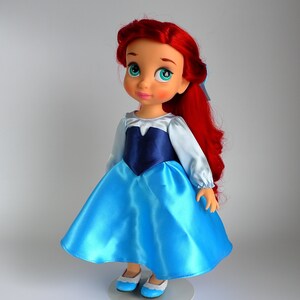 May include: A red-haired doll wearing a blue and white dress with blue shoes. The doll has a light blue top and a dark blue bodice. The doll is standing on a white stand.