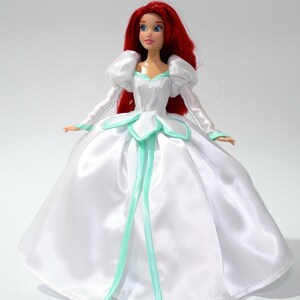 May include: A red-haired doll wearing a white satin dress with a green trim. The dress has a sweetheart neckline and puffy sleeves. The doll is standing on a white background.