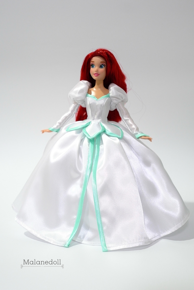 May include: A red-haired doll wearing a white satin dress with a green trim. The dress has a sweetheart neckline and puffy sleeves. The doll is standing on a white background.