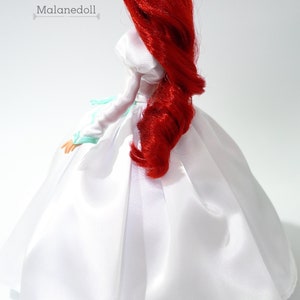 May include: A red-haired doll wearing a white satin dress with a light blue sash. The doll's hair is styled in a long, flowing curl.
