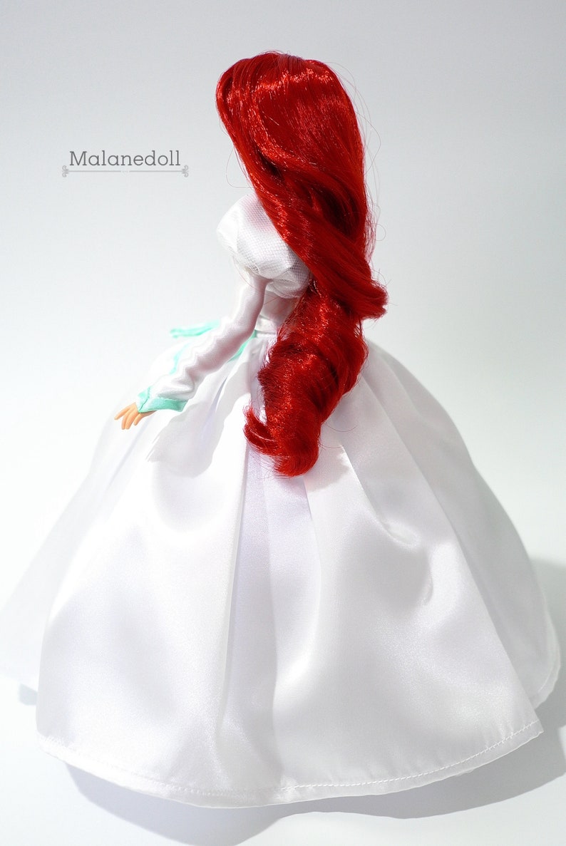 May include: A red-haired doll wearing a white satin dress with a light blue sash. The doll's hair is styled in a long, flowing curl.